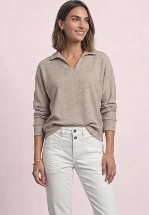Kvinde med skulderlangt brunt hår smiler iført en beige langærmet sweater og højtaljede hvide bukser mod en ensartet lys baggrund.