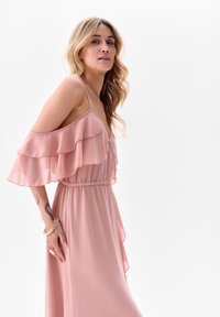 Roze chiffon jurk met gelaagde ruffle-mouwen, fitted elastische taille en vloeiende silhoutte. Heeft een zijsplit en een soepele textuur.