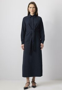 Rochie cămașă în dungi bleu-marinar, cu mâneci lungi, guler cu butoane și un detaliu legat în față. Prezintă o croială lejeră și un tiv până la glezne. Design elegant și sofisticat.