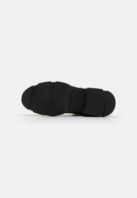 Semelle de chaussure en caoutchouc noir avec surface texturée, design segmenté et bout arrondi. Présente cinq rainures horizontales à travers la partie médiane.