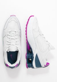 Bílé sportovní boty s vrchní částí z prodyšné síťoviny, lemované fialovými akcenty. Polstrovaná podrážka má průhledný modro-fialový podpatek.