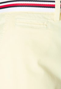 Tissu jaune clair avec une texture lisse, comportant une poche au dos et une taille avec des rayures noires, rouges et blanches.