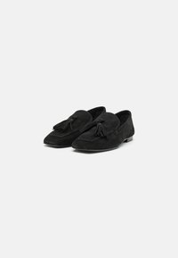 Mocassins de camurça pretos com detalhes em franjas, apresentando um bico afilado e sola plana. A textura é suave, com um interior liso.