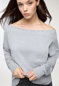Maglione grigio chiaro off-shoulder con texture morbida, caratterizzato da dettagli metallici lungo lo scollo e polsini a coste. Vestibilità morbida in vita.