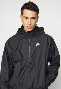 Melna vējjaka ar pusi jostu priekšā, regulējamu kapuci un elastīgiem aprocēm. Uz krūtīm ir neliels balts Nike logo.