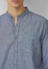 Heren overhemd met knopen in lichtblauwe chambray stof, met een mandarin kraag, een borstzak en houten knopen aan de voorkant.