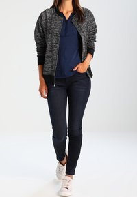 Zwarte en grijze textuur bomberjack, marineblauwe polo, donkere jeans en lichte sneakers. Het jack heeft ribbelmanchetten en een ritssluiting aan de voorkant.