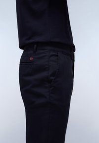 Marine chinos med en glat tekstur, der har en sidelomme og et lille rødt og hvidt broderet flagdetalje på baglommen.