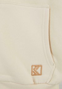 Krèmfarget hettegenser med frontlomme, ribbestrikkede ermer og et brodert merke i lys beige nyanse. Glatt, myk tekstur.