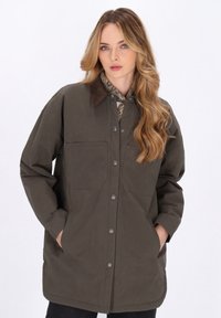 Giacca verde oliva con colletto a camicia, due tasche frontali e bottoni metallici. Il tessuto appare morbido con una finitura leggermente texturizzata.