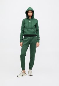 Camisola com capuz verde escuro e calças de treino combinando, feitas de tecido macio, com bolso frontal e logótipo da Nike. Usadas com ténis desportivos de cor clara.