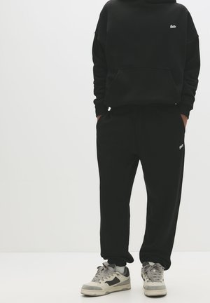 Black hoodie with front pocket and small logo, paired with matching black sweatpants. Gray and white sneakers with laces and accents.