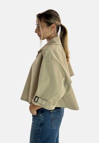 Giacca beige corta con vestibilità oversize, colletto con bottoni e fibbia regolabile sulle maniche. Indossata su jeans blu, con una texture del tessuto liscia.