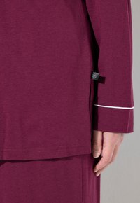 Maglietta a maniche lunghe maroon con una texture liscia, piping bianco sui polsini e un'etichetta laterale vicino alla cucitura.