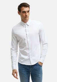Weißes, langärmliges Hemd mit klassischem Kragen, aus weichem Stoff, kombiniert mit blauen Denim-Jeans. Schlichtes Design, ohne Muster.