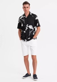 Camisa negra de manga corta con un patrón abstracto blanco, combinada con pantalones cortos blancos y zapatillas oscuras. El modelo está de pie con las manos en los bolsillos.