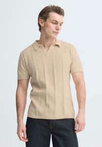 Man draagt een beige korte mouw gebreide poloshirt en donkere spijkerbroek, kijkt naar beneden naar zijn linkerzijde met een neutrale achtergrond.