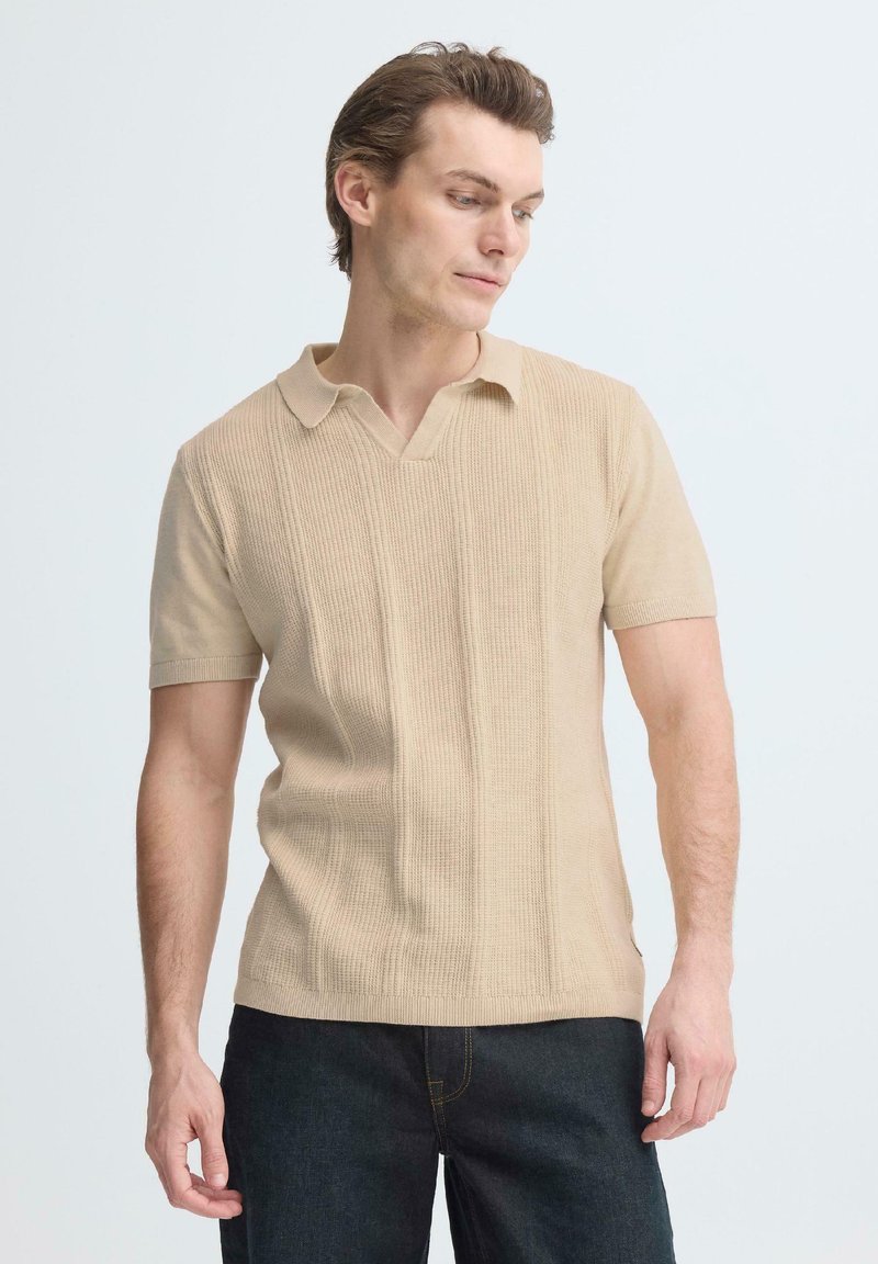 Man draagt een beige korte mouw gebreide poloshirt en donkere spijkerbroek, kijkt naar beneden naar zijn linkerzijde met een neutrale achtergrond.