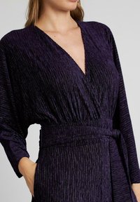 Vestido morado texturizado con un pronunciado escote en V y mangas largas, que presenta una cintura atada y plisados verticales que realzan el brillo de la tela.