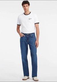 Λευκό μπλουζάκι με μαύρες λεπτομέρειες και λογότυπο "Guess", συνδυασμένο με μπλε τζιν παντελόνι ίσιου κόψιμου και μπεζ αθλητικά παπούτσια. Απλός σχεδιασμός και κλασική εφαρμογή.