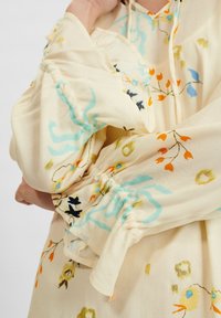 Hedvábná blůza s květinovým vzorem v jemně žluté barvě, se staženými manžetami a barevným výšivkem s modrými, oranžovými, zelenými a černými akcenty.