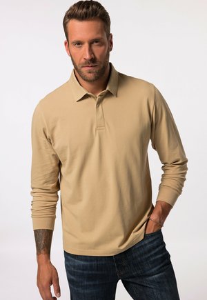 Beige long-sleeve polo shirt with a collar, made from smooth fabric. Model wearing dark jeans, showing a tattoo on left wrist.