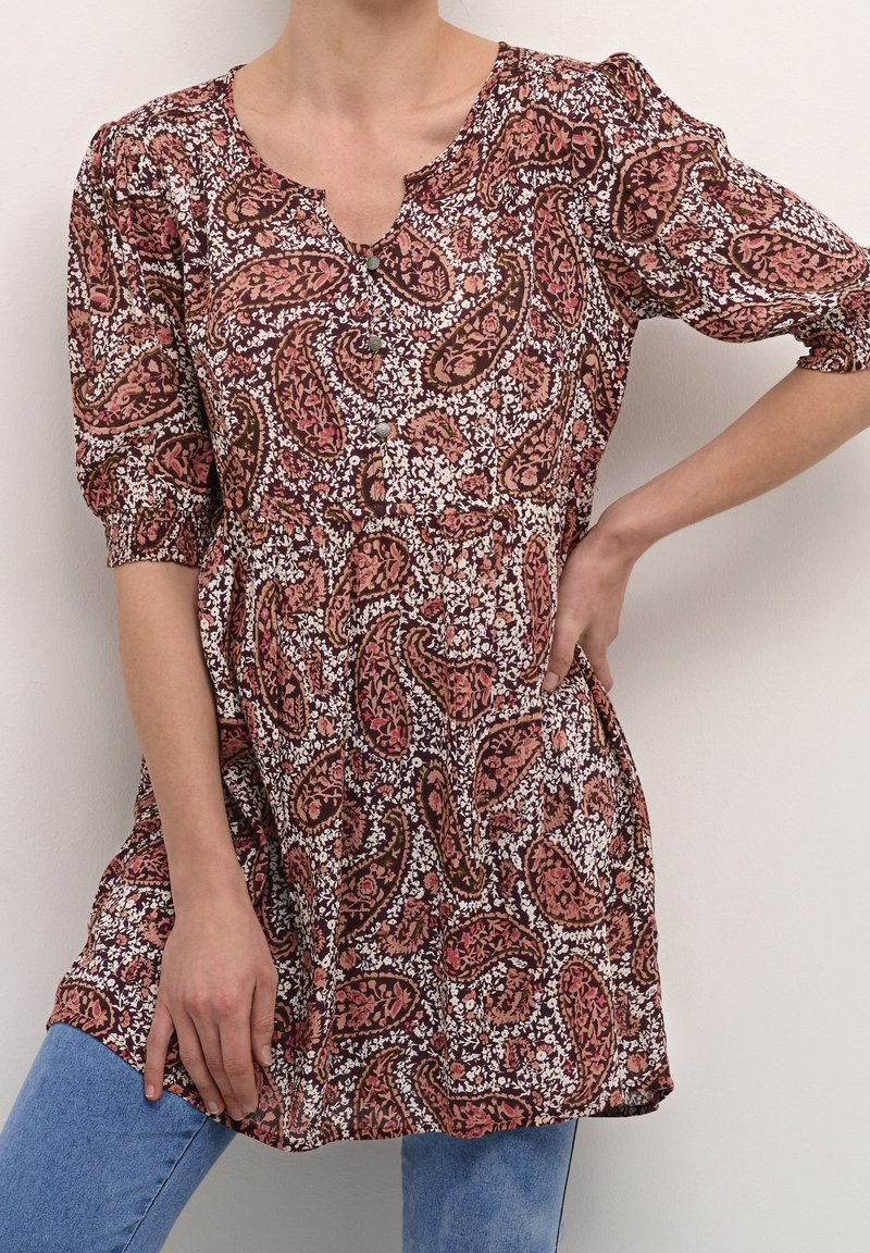 Femme portant un chemisier marron et rose à motif floral paisley avec des manches bouffantes trois-quarts et un jean bleu, debout avec une main sur la hanche.