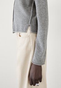 Grå stickad tröja med sidoflikar, tillsammans med ljusgul-beiga byxor. Närbild av ärm och hand med en metallring.