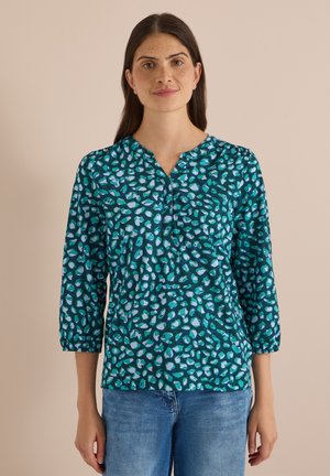 Femme aux longs cheveux foncés portant un chemisier manches longues à motifs bleu sarcelle et bleu, et un jean bleu, debout devant un fond beige uni.