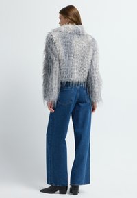 Grau gefütterte, kurze Jacke aus Kunstfell mit fransiger Textur, getragen über blauen hochtaillierten, weiten Jeans und schwarzen Stiefeln bis zum Knöchel.