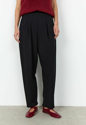 Persona vistiendo pantalones negros plisados de cintura alta, suéter polar rojo con cremallera y zapatos planos rojos, de pie sobre un suelo gris contra una pared blanca.