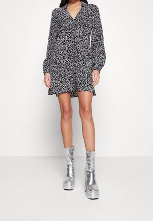 Czarna sukienka w białe wzory z długimi rękawami, dekoltem w serek i krótką długością, zestawiona z błyszczącymi srebrnymi butami na platformie.