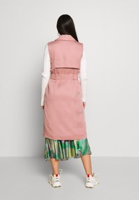 Růžový kabát bez rukávů s vytaženým pasem, vyrobený z měkké látky, vrstvený přes zelenou květinovou sukni s volným splýváním.