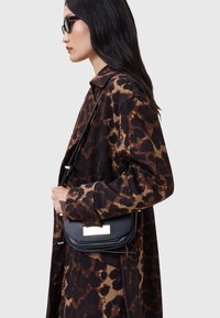 Borsa a tracolla in pelle nera con hardware dorato, texture liscia e chiusura a pattina. Indossata a tracolla; abbinata a un cappotto con stampa leopardo marrone.