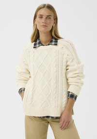Pull en tricot torsadé crème avec des motifs en losange, poignets et ourlet côtelés, superposé sur une chemise à col à carreaux sombres. Texture douce. Coupe décontractée.