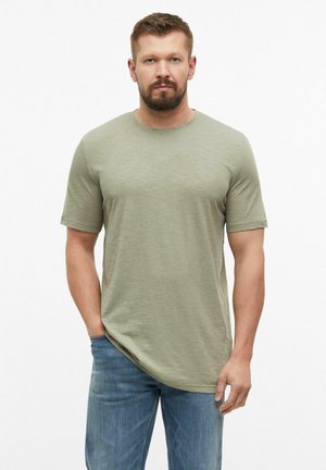 Mann mit kurzen braunen Haaren und Bart, der ein hellgrünes T-Shirt mit kurzen Ärmeln und blaue Jeans trägt, steht mit einer Hand in der Tasche.