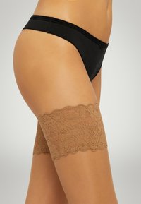 Nude Spitzenstrumpfhalter bis zum Oberschenkel mit einem gewellten oberen Rand, komplizierten Mustern und einer glatten Oberfläche, kombiniert mit einem schwarzen Bikini.