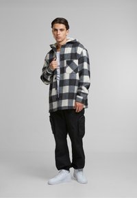 Camicia di flanella a scacchi bianco e nero con cappuccio, dotata di taschino sul petto; abbinata a pantaloni cargo neri e sneakers bianche.