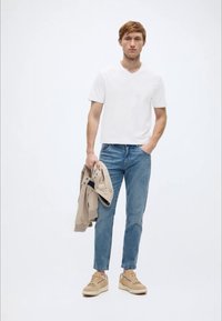 Λευκό T-shirt με λαιμόκοψη V, ελαφρύ μπεζ μπουφάν κρατημένο στο ένα χέρι, μπλε στενές τζιν παντελόνες και μπεζ αθλητικά παπούτσια με υφασμένα πέλματα. Απλός σχεδιασμός σε όλο το σύνολο.