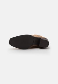 Tan leather shoe with a textured upper, a low block heel, and a smooth black rubber sole. Features a small logo detail on the side.