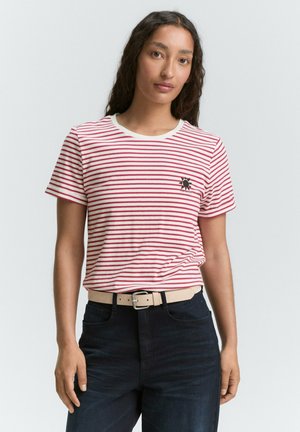 Randig röd och vit t-shirt med rundad halsringning och korta ärmar, prydd med en svart sol-logga på vänster bröst. Bärs med mörka jeans och ett beige bälte.