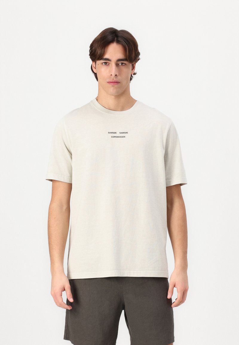 Camiseta de manga corta beige claro hecha de algodón suave, con una etiqueta impresa minimalista en negro en el centro.