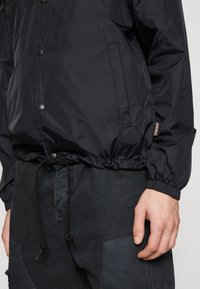 Ingrandimento di una persona che indossa una giacca nera con bottoni, maniche elastiche e coulisse regolabile, abbinata a pantaloni scuri.