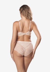 Beżowy biustonosz z regulowanymi ramiączkami i zapięciem na haczyk, w połączeniu z przezroczystymi, koronkowymi majtkami z kwiatowymi wzorami.