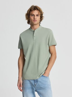 Giovane uomo con capelli ricci che indossa una maglietta henley verde chiaro a maniche corte e jeans azzurri, in piedi con una mano in tasca.