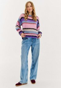 Mnohobarevný pletený svetr s růžovými, fialovými a krémovými pruhy, kombinovaný se světle modrými širokými džíny a burgunovými botami.