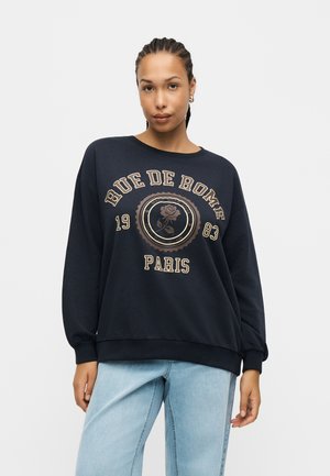 Kvinde med fletninger bærer en sort "Rue de Rome Paris" sweatshirt med et rose-motiv, kombineret med lyseblå jeans, mod en ensfarvet baggrund.