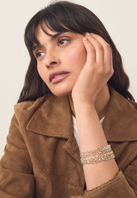 Layered crystal bracelet with metallic accents, displaying clear and reflective stones. Model wearing a brown suede jacket in a neutral setting.