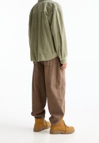 Camisa de pana verde oliva, pantalones sueltos marrones y botas amarillas con cordones. La camisa tiene un cuello abotonado y una textura acanalada.