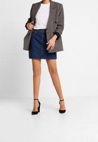 Blazer oversize à carreaux gris, t-shirt blanc, mini-jupe en denim foncé et chaussures à talons noires, affichant une tenue décontractée en couches.
